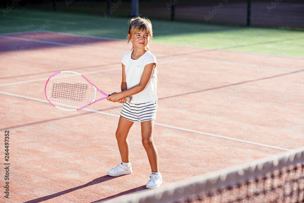 Female little tennis player in white sport uniform practice in hitting with tennis racket at the training on outdoor court on the sunset background.