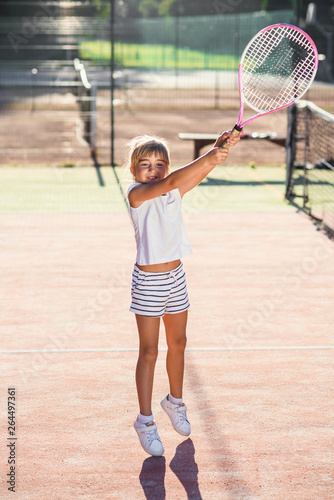 Female little tennis player in white sport uniform practice in hitting with tennis racket at the training on outdoor court on the sunset background. © gorynvd