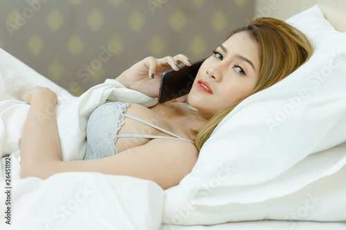 Young beautiful woman with long brown hair uses cell phone and relaxes on the white background bed.