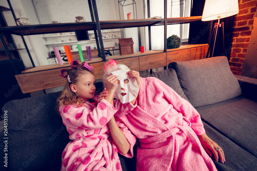 Caring cute girl putting moisturizing mask on face of granny