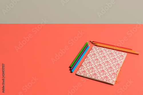 Fondo coral vivo color del 2019 con cuaderno con patron renacentista en rojo burgundy y beige acompañado de lapices de colores naranja, rosa y rojo