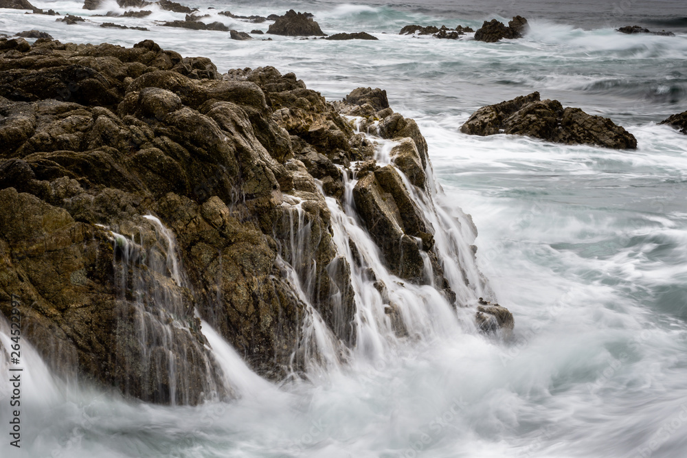 Long exposure of Pacific ocean waves running back after crashing over a stretch of rocky coastline, creating many waterfalls cascading off the rock back into the sea. 