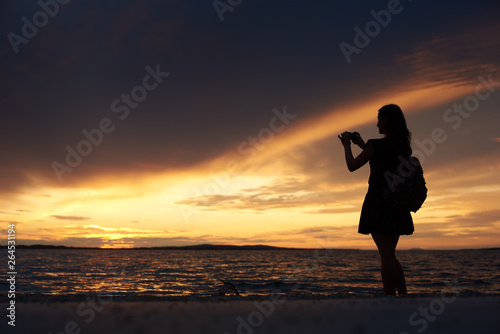 Silhouette of slim woman in short dress and with backpack and camera standing alone at water edge taking picture of beautiful seascape at sunset. Tourism and vacations concept.