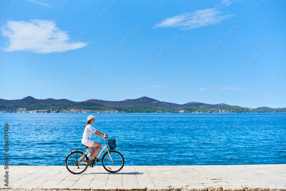Obraz premium Young woman in white summer closing and hat riding a bicycle along stony sidewalk on blue sparkling sea water and resort town at foot of mountains on opposite shore background. Tourism and vacations.