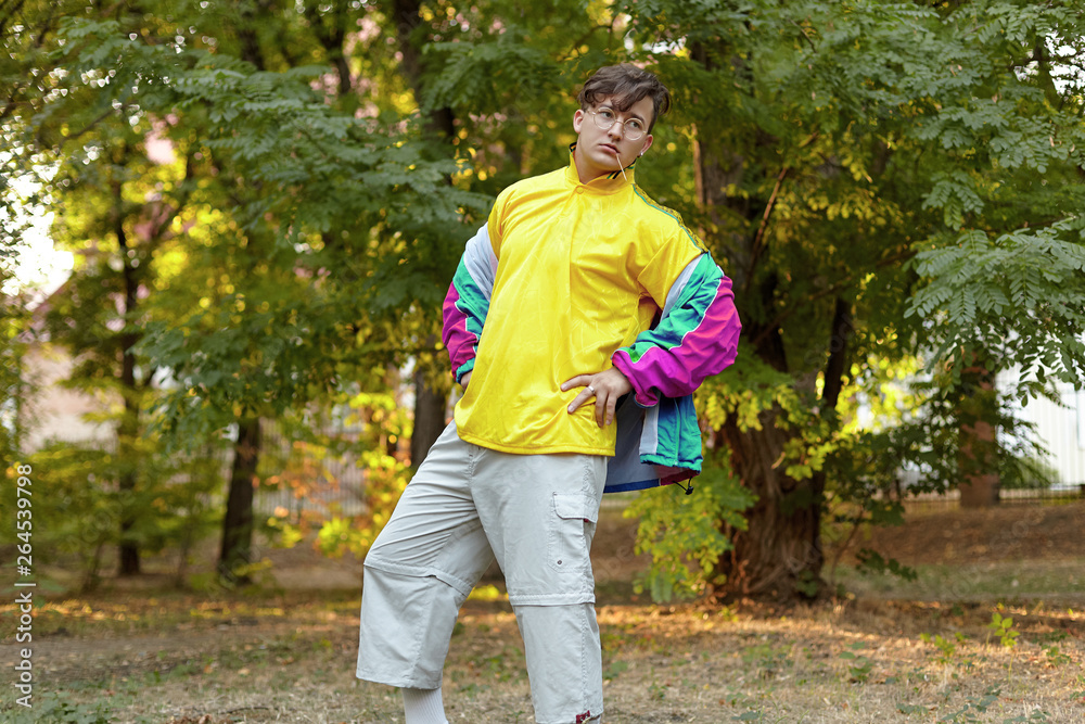 Young handsome caucasian man with bright freaky sportswear 70s style stands  at a morning park. Stick in the mouth, golden glasses, earring and ring.  Extravagance fashion. Outdoors, copy space. Stock Photo
