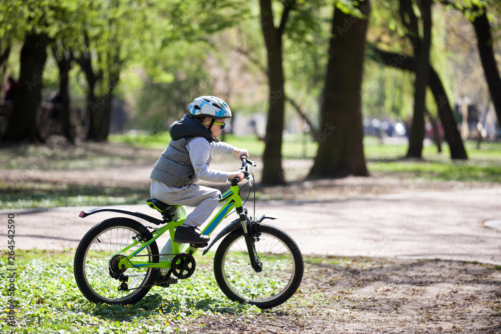 Fototapeta premium Little boy learns to ride a bike in the park. Cute boy in sunglasses rides a bike. Happy smiling child in helmet riding a cycling.