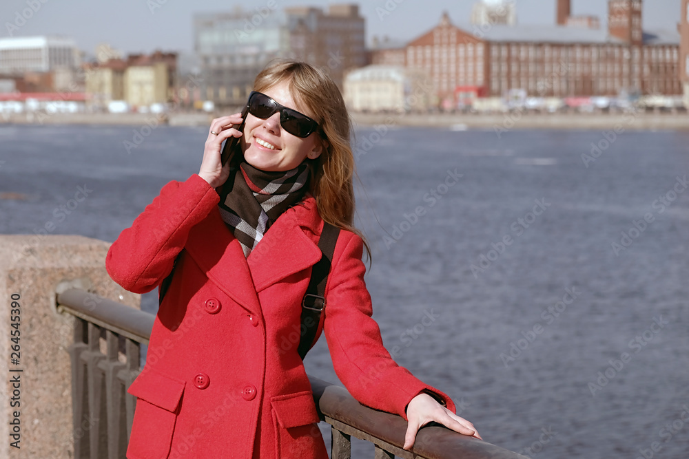 Fototapeta premium A young girl stands on the waterfront and talking on a mobile phone. Woman happily talking to her friends on the phone and smiling