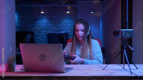 Closeup shoot of young attractive female blogger with dreadlocks in headphones playing video games on the laptop with the neon background indoors