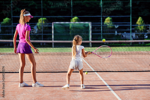 Little girl in white uniform hitting ball under net during tennis training with coach. Attractive female coach making exercises with little tennis player on the outdoor court at sunset.