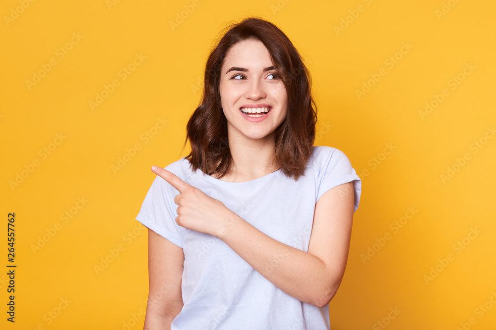 Fototapeta premium Smiling beautiful young woman wears white casual t shiort, points with index finger aside, presenting something and looking away, model poses isolated on yellow background. Copy space for advertisment
