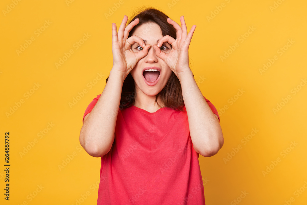 Image of young happy woman in red t shirt, holding her hand over her eyes as glasses and looking through fingers, having fun while being photographed in studio, isolated over yellow background.