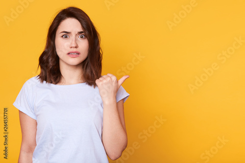 Close up portrait of astound woman has shocking facial expressian  points wityh thumb on right empty corner  wears white t shirt  shows something surprised. Copy space for advertisment or promotion.