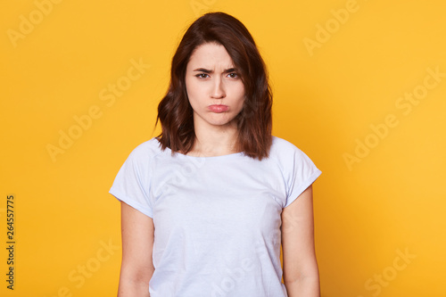 Portrait of beautiful dark haired young woman in casual white t shirt, standing and looking directly at camera, posing with pouty lips isolated on yellow background, has offended facial expression.