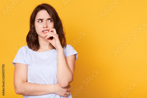 Horizontal portrait of thoughtful female looks with pensive dreamy expression, looking aside, keeps hand near face, thinks what decision to make, stands against yellow background