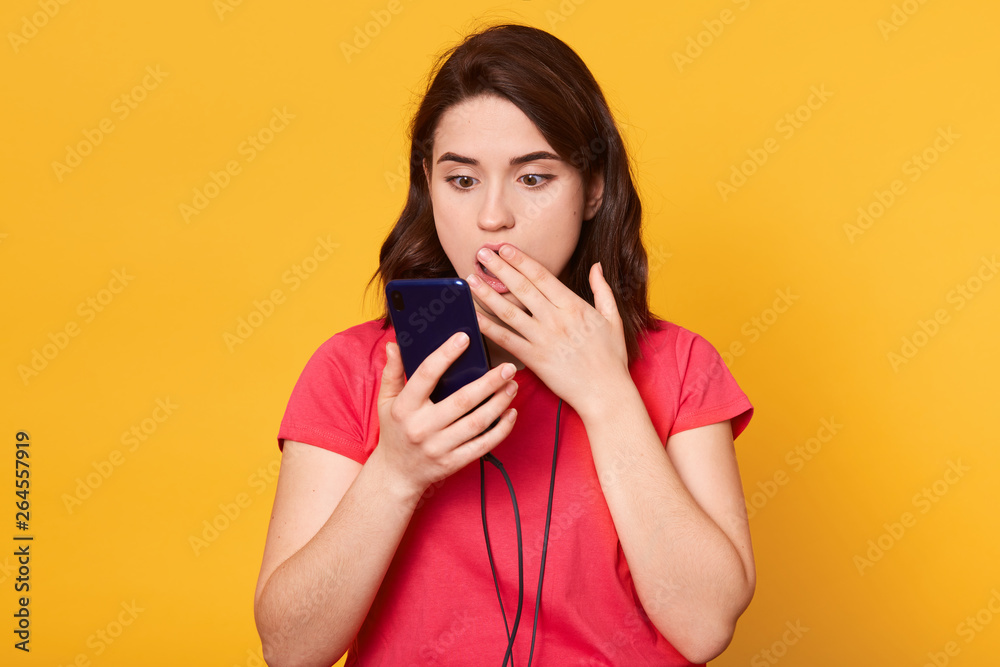 Close up portrait of shocked woman talking to somebody, has video call, uses modern smartphone, dressed red t shirt, covers mouth with hand, hears surprised news, has astonish facial expression. Close up portrait of shocked woman talking to somebody, has video call, uses modern smartphone, dressed red t shirt, covers mouth with hand, hears surprised news, has astonish facial expression.