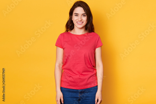 Smiling joyfull female with dark hair, dressed casually, looking with satisfaction directly at camera, being in good mood, shot of good looking beautiful woman isolated against blank studio wall.