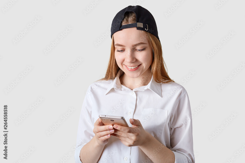 Close up portrait of smiling beautiful teenager with visor cap back, has brown hair, poses at white studio wall, holds smartphone, checking social network, comunicates with friens, wears white blouse. Close up portrait of smiling beautiful teenager with visor cap back, has brown hair, poses at white studio wall, holds smartphone, checking social network, comunicates with friens, wears white blouse.