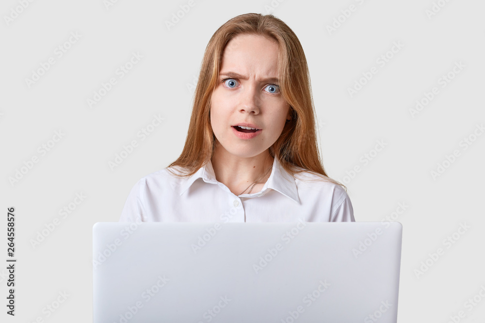 Attractive girl looks at camera with opened mouth, wide opened blue eyes, has straight broun hair, dressed white shirt, working online, shocked with new task, looks astonished, isolated on studio wall Attractive girl looks at camera with opened mouth, wide opened blue eyes, has straight broun hair, dressed white shirt, working online, shocked with new task, looks astonished, isolated on studio wall
