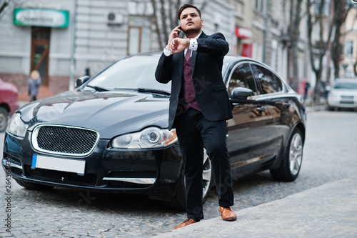 Stylish indian businessman in formal wear with mobile phone standing against black business car on street of city. Speak on cellphone and looking at his watches.