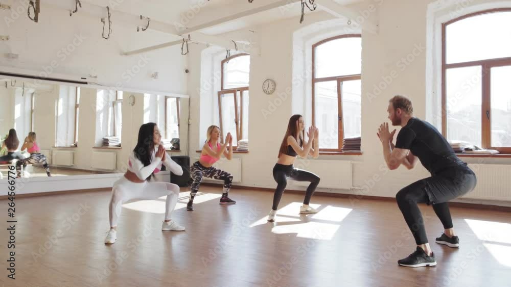 Tracking right wide shot of male fitness instructor and three young women doing squat exercise together during small group class