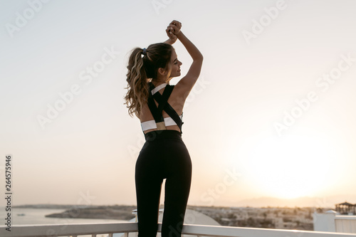 Photography Beautiful young woman in sportswear with long curly hair stretching and smiling on seafront on sunrise background