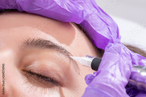 Cosmetologist making permanent make up on eyebrows. Tattooist with purple gloves and professional instrument applying tattoo in beauty studio on beautiful girl. Close up  selective focus