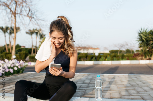 Amazing joyful pretty young woman in sportswear, with long curly hair chatting on phone outside in park in the morning. Workout, cheerful mood, happiness, sunrise, joy, healthy lifestyle