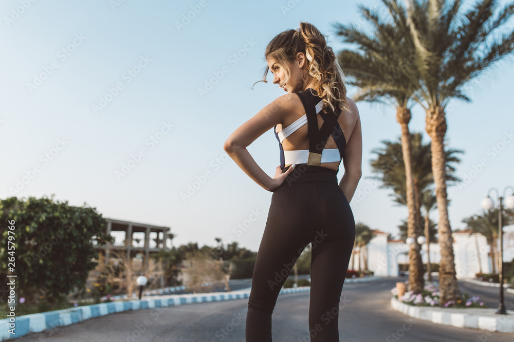 Charming attractive young woman at training in sunny morning on street with palm trees in tropical city. View from back, outwork, sportive lifestyle. Place for text