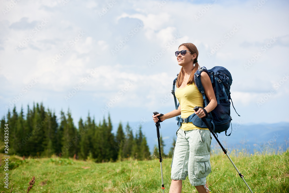 Young smiling woman hiker hiking mountain trail, walking on grassy hill, wearing backpack and sunglasses, using trekking sticks, enjoying summer cloudy day. Outdoor activity, tourism concept