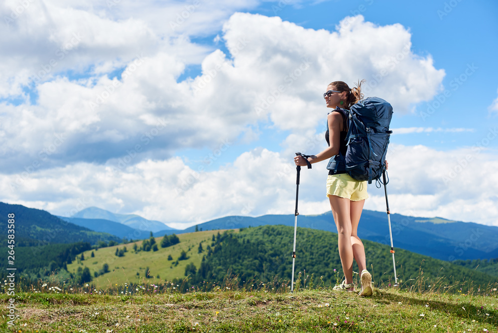 Athlete smiling woman tourist hiking mountain trail, walking on grassy hill, wearing backpack and sunglasses, using trekking sticks, enjoying summer sunny day in the mountains. Tourism concept