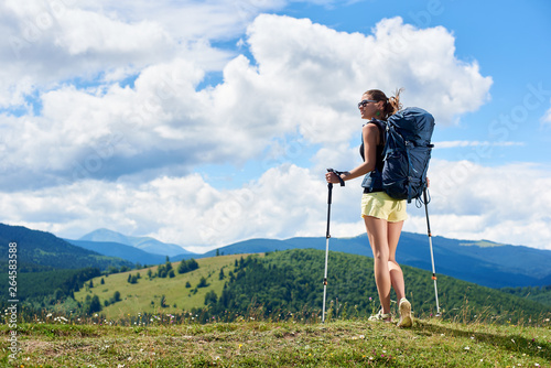 Athlete smiling woman tourist hiking mountain trail, walking on grassy hill, wearing backpack and sunglasses, using trekking sticks, enjoying summer sunny day in the mountains. Tourism concept