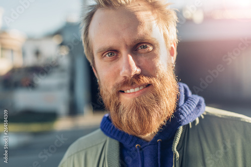 Portrait of cheerful nice young bearded man outside on street. He look on camera and smile. Sun shining behind. Sunlight. Urban view.