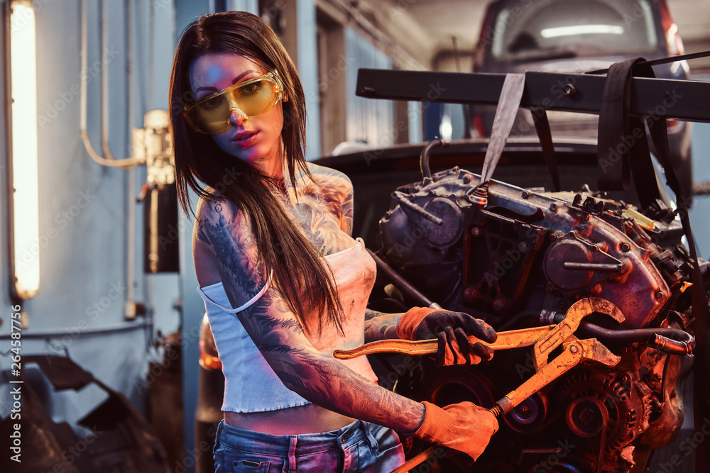 Stylish girl with tattooed body wearing protective goggles working with a car engine suspended on a hydraulic hoist in the workshop. Photo with red light illumination