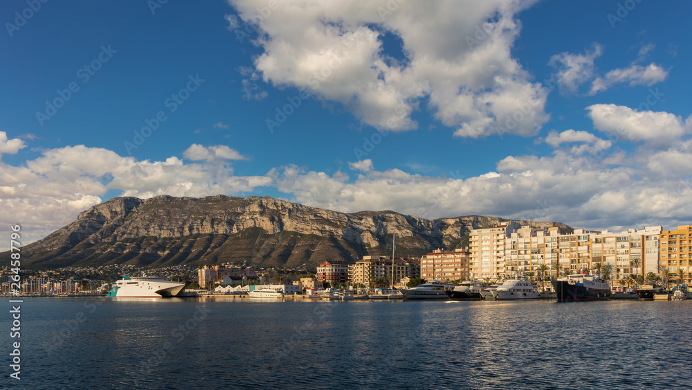 View from the harbor on the skyline of Denia with many ships, ferry and the mountain Montgo in the background in clear weather with blue sky and light clouds.