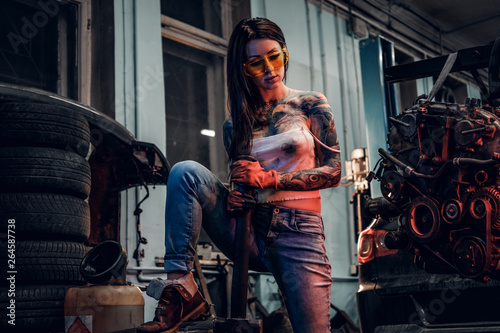 Female car mechanic holding a big hammer and posing next to a car engine suspended on a hydraulic hoist in the workshop.