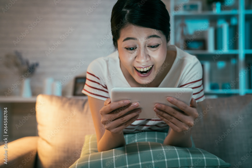 happy teenage asian girl sitting on couch at home enjoy funny movie on  tablet in dark