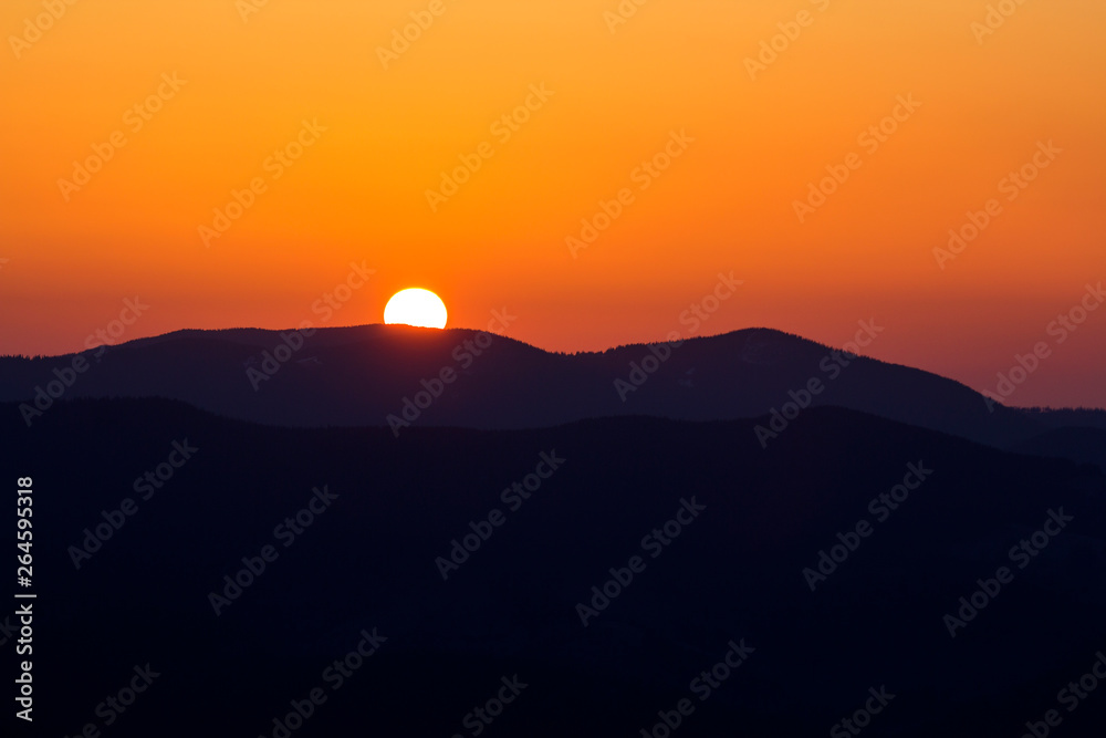 Beautiful sunset in mountains. Wide panorama view of big bright white sun in dramatic orange sky over dark mountain range landscape at sunset or sunrise. Beauty of nature concept.