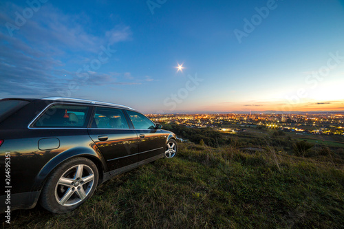 Black car parked at night in green meadows on copy space background of lights of distant city buildings and bright blue sky with first star at sunset.