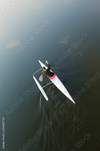 Adult Caucasian woman viewed from above and behind paddling in white outrigger canoe with float on dark water background with copy space