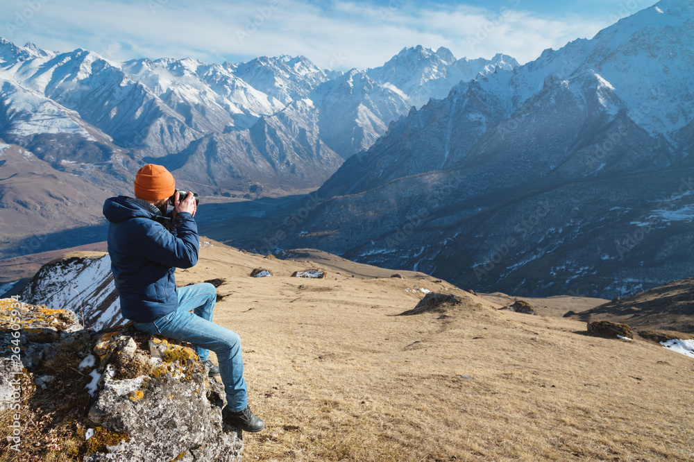 Portrait of a bearded male photographer in sunglasses and a warm jacket with a backpack sits on a big stone and takes pictures on camera against the background of snow-capped mountains Portrait of a bearded male photographer in sunglasses and a warm jacket with a backpack sits on a big stone and takes pictures on camera against the background of snow-capped mountains
