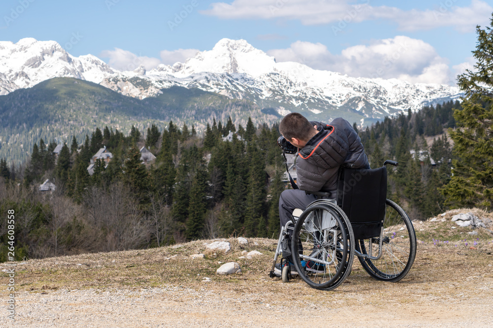 Disabled man on wheelchair using camera in nature, photographing beautiful mountains
