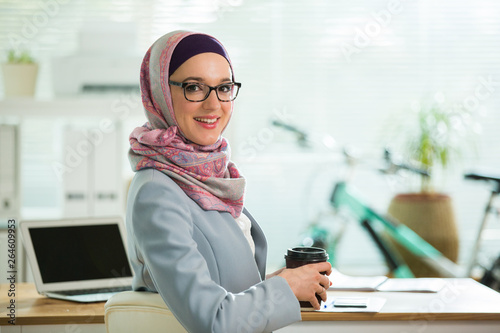 Beautiful stylish woman in hijab and eyeglasses, sitting at desk with laptop in office. Portrait of confident muslim businesswoman. Modern office with big window, bicycle on background. 
