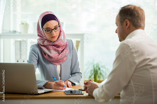 Coworkers meeting in office. Stylish woman in hijab making conversation at desk with man in white modern office. Muslim businesswoman in eyeglasses interviewing man.