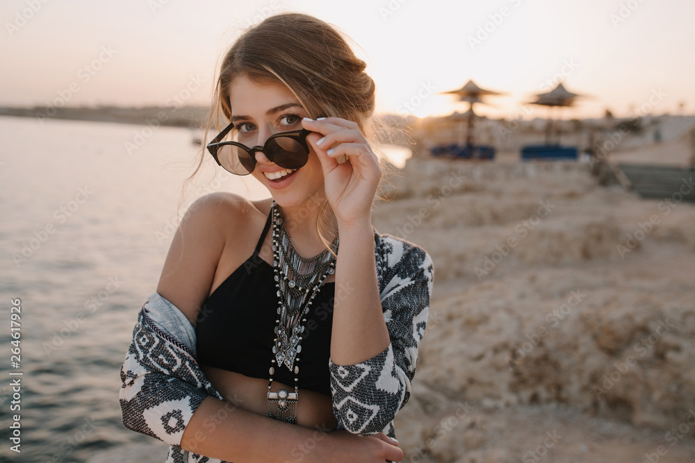 Closeup portrait of attractive young girl taking off stylish sunglasses, on sunset, on beach with sensual look. Wearing trendy black top, necklace, cardigan with ornaments. Resort, vacation, holiday.