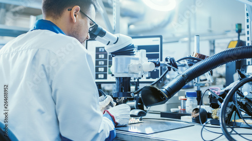Tableau sur toile Electronics Factory Worker in White Work Coat is Soldering a Printed Circuit Board Through a Digital Microscope