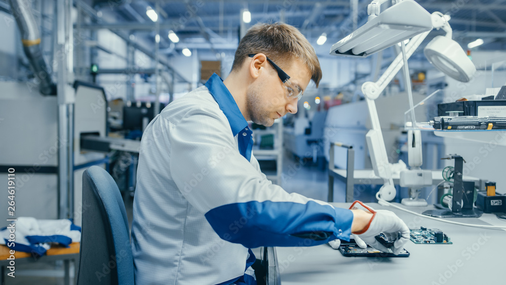 Young Man in Blue and White Work Coat is Using Plier to Assemble Printed Circuit Board for Smartphone. Electronics Factory Workers in a High Tech Factory Facility.