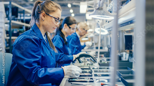 Female Electronics Factory Workers in Blue Work Coat and Protective Glasses Assembling Printed Circuit Boards for Smartphones with Tweezers. High Tech Factory with more Employees in the Background.  photo