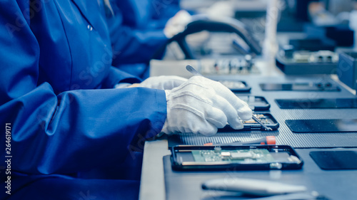 Close Up of a Female Electronics Factory Worker in Blue Work Coat and Protective Glasses Assembling Smartphones with Screwdriver. High Tech Factory Facility with more Employees in the Background. 