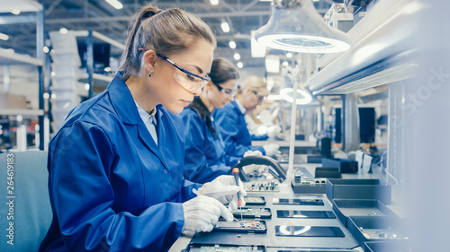 Woman Electronics Factory Worker in Blue Work Coat and Protective Glasses is Assembling Smartphones with Screwdriver. High Tech Factory Facility with more Employees in the Background.  photo
