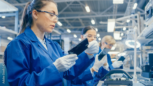 Female Electronics Factory Worker in Blue Work Coat and Protective Glasses Takes a Smartphone Screen and Performs a Quality Check. High Tech Factory Facility with Multiple Employees. 